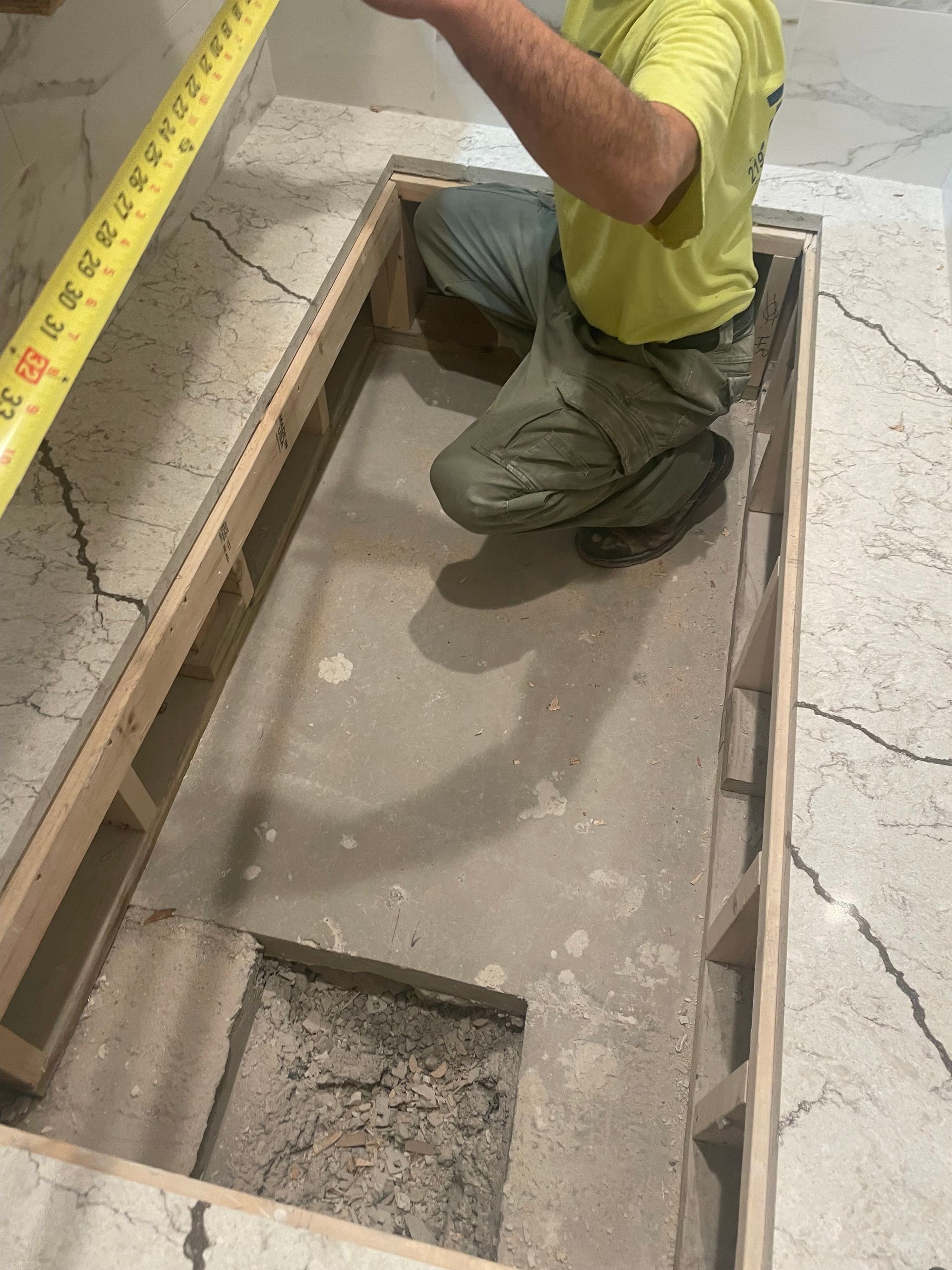 Person kneeling, measuring a rectangular floor opening with a tape measure. Wooden frame surrounds the opening.