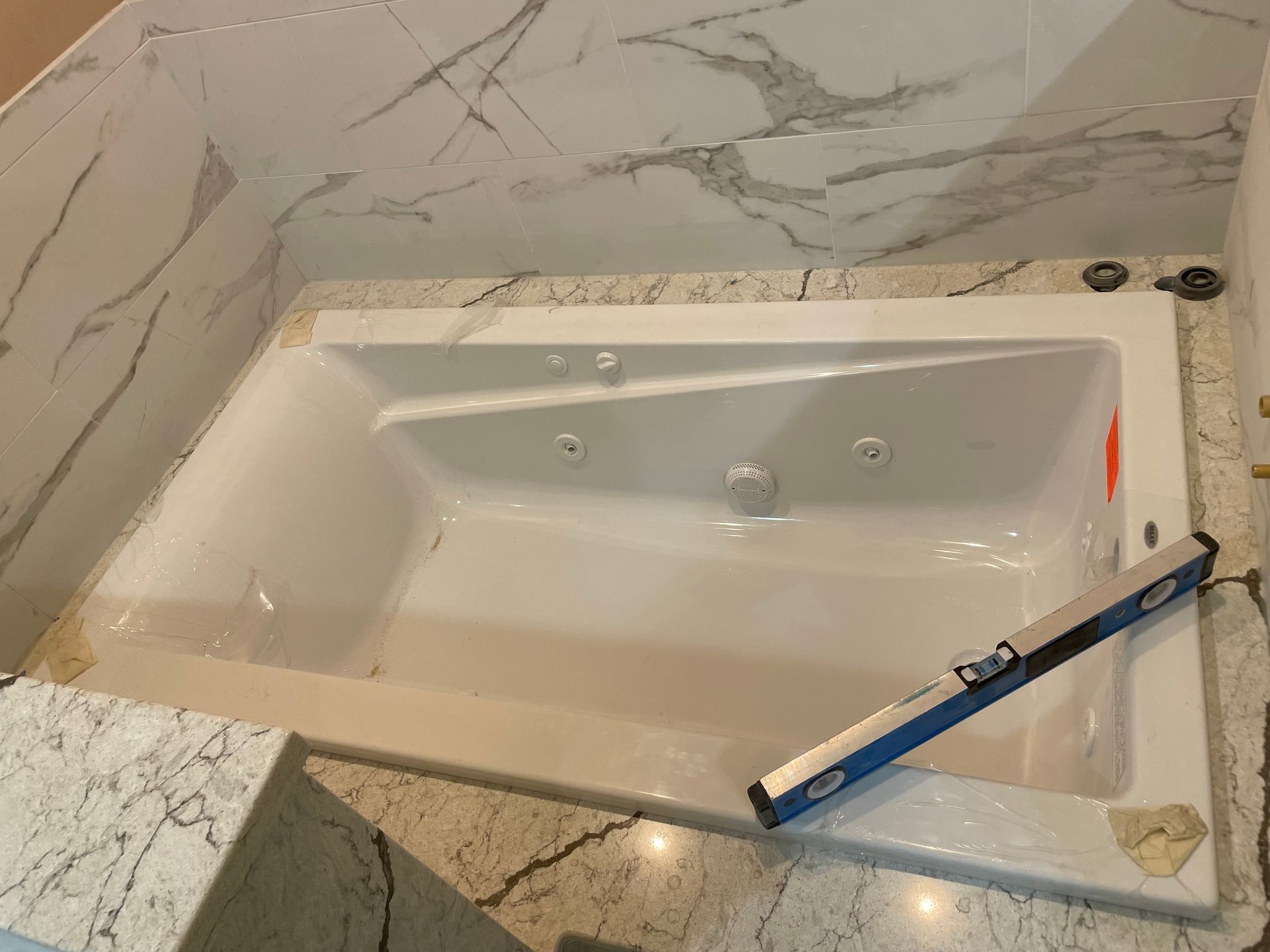 White jacuzzi tub built into a marble-tiled bathroom. A level rests on the tub's edge.