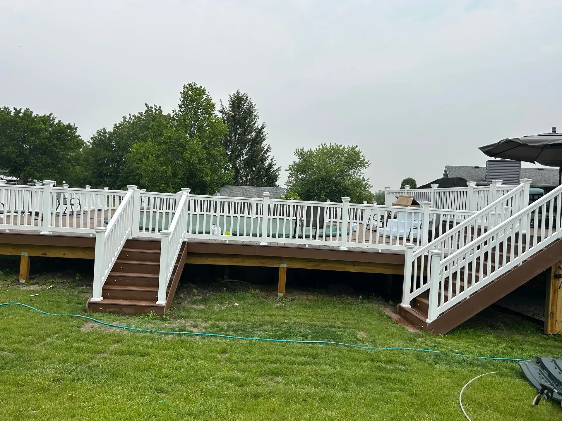Wooden deck with white railings, stairs, and pool area on green lawn. Overcast sky.