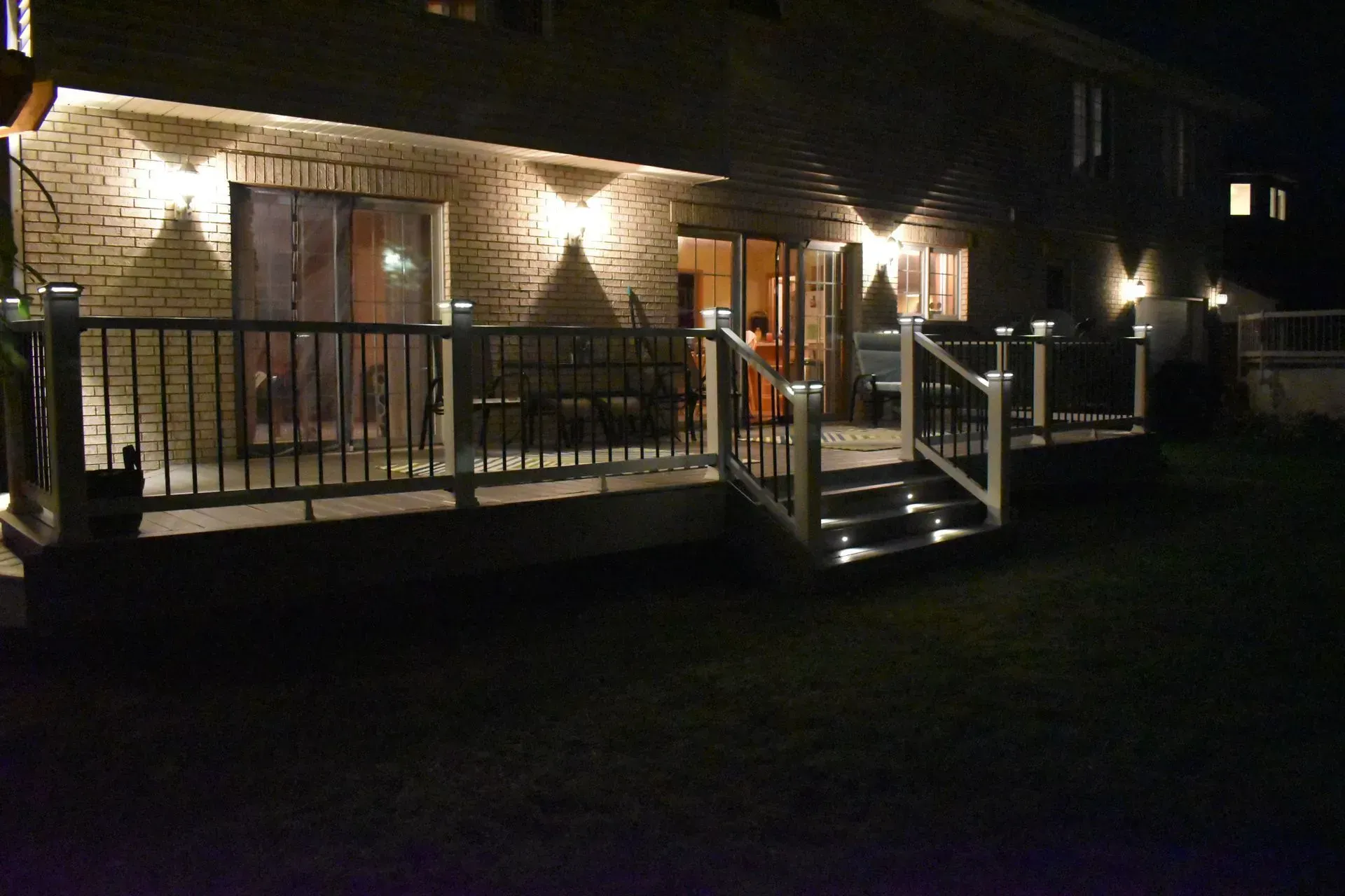 Backyard deck at night, lit by built-in lights. House with sliding doors and brick facade. Grass lawn in foreground.