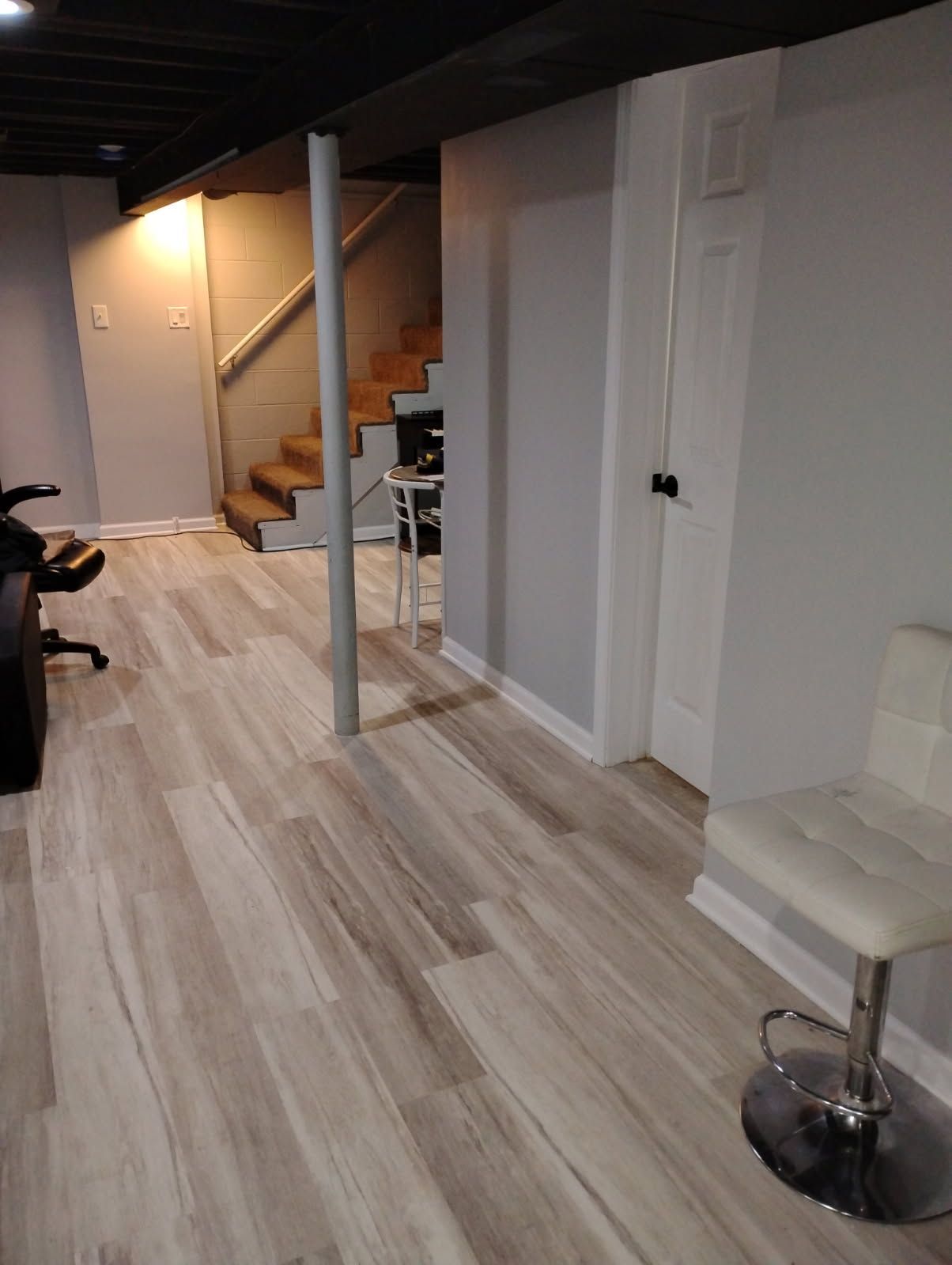Basement with light gray walls, wooden stairs, and wood-look flooring. White chair is to the right.
