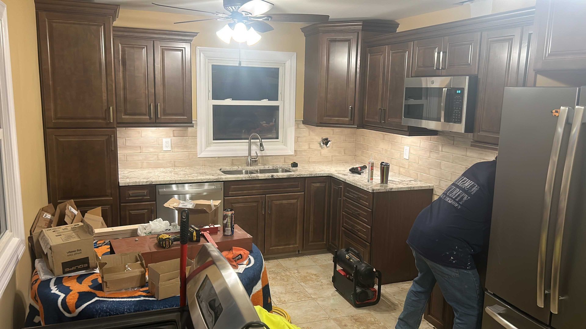 Kitchen remodel in progress with dark brown cabinets, granite countertops, and a worker by the refrigerator.