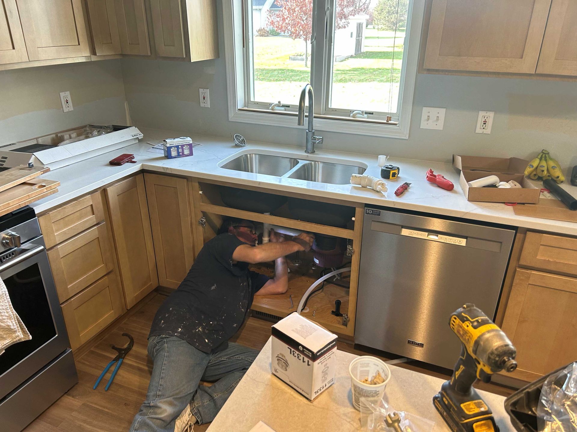 A plumber works under a kitchen sink, installing plumbing. Light wood cabinets, white countertop, and a stainless steel dishwasher are visible.