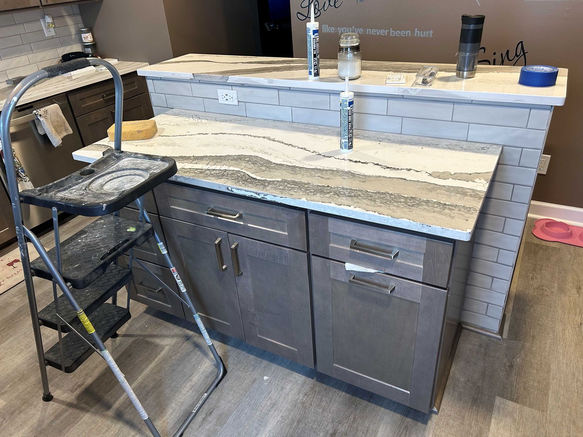 Kitchen renovation: Countertop installation on gray cabinets, step stool, caulk, gray and white stone.