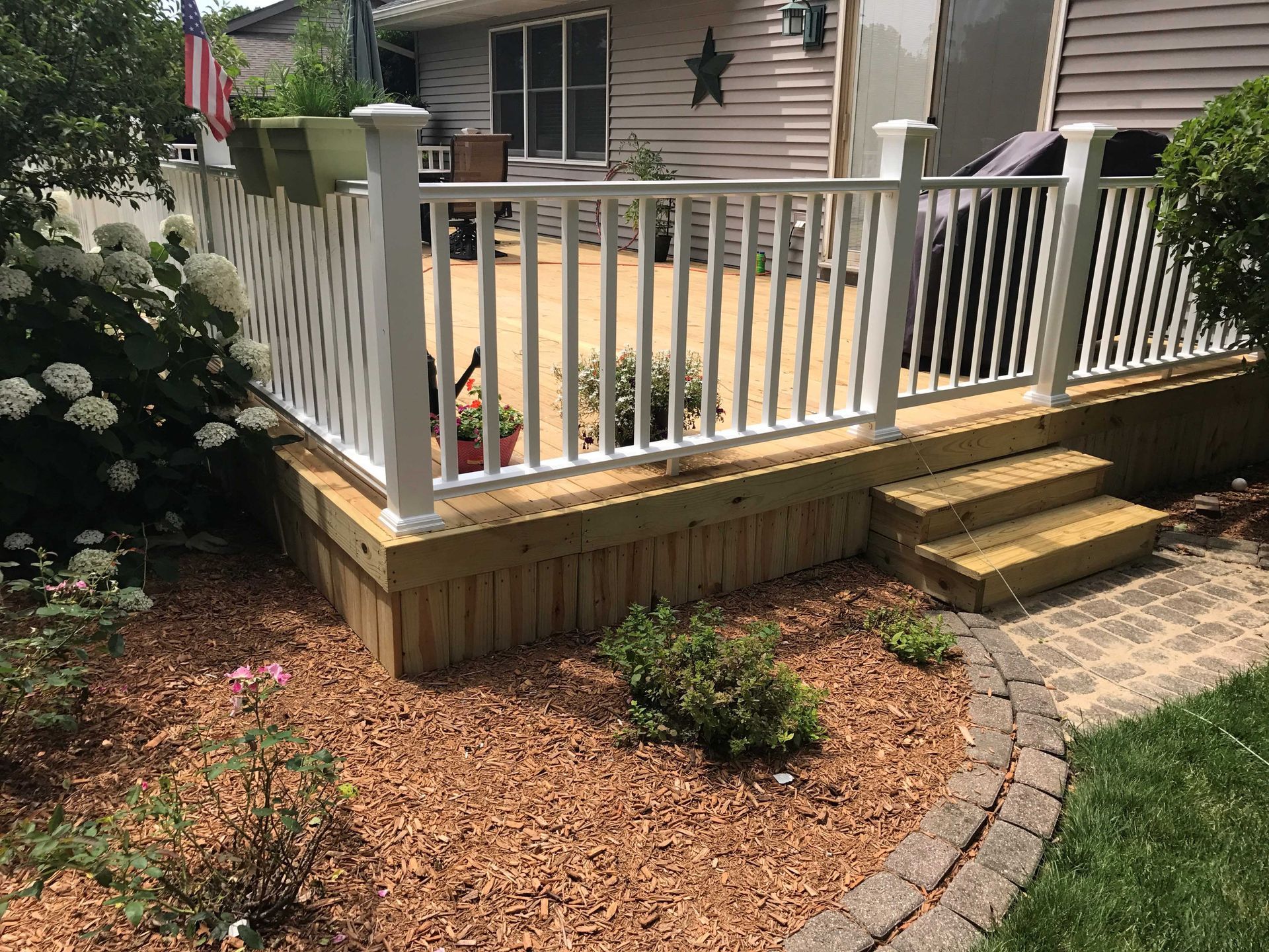 A wooden deck with white railing, steps, and landscaping in a backyard.
