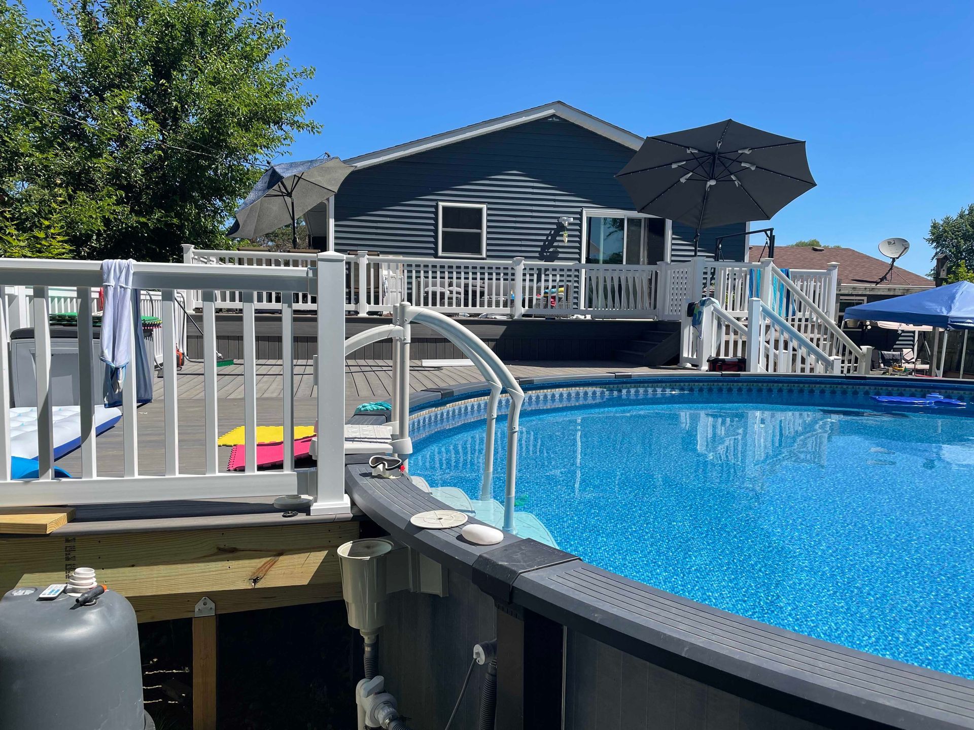 Above-ground pool with a deck, backyard setting with a house in the background, and a bright blue sky.