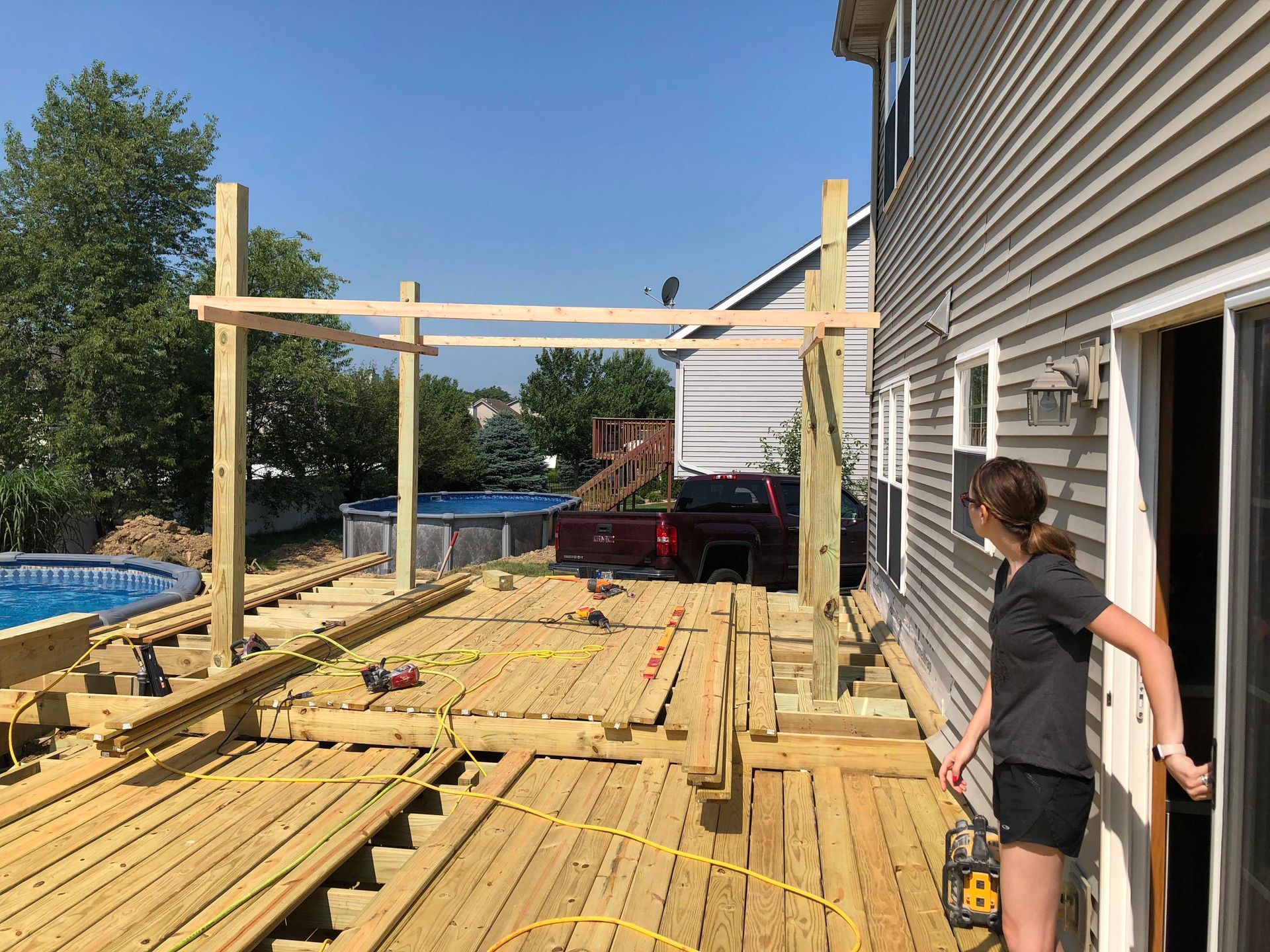 Deck construction with overhead frame; woman opens door. Pool and truck visible. Sunny, exterior.