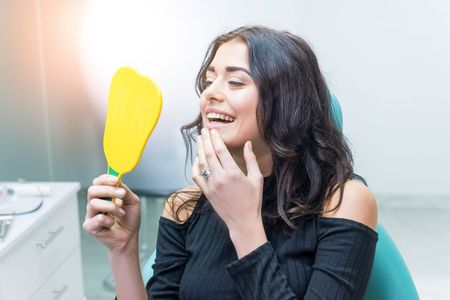 A woman is sitting in a dental chair looking at her teeth in a mirror.