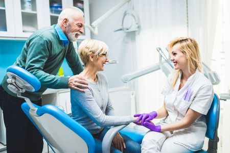 An elderly couple is sitting in a dental chair talking to a dentist.