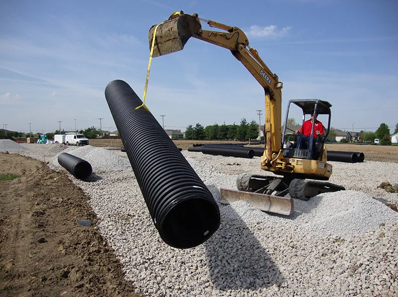A yellow excavator on a gravel lot lifts a large, black, corrugated plastic pipe using a yellow strap.