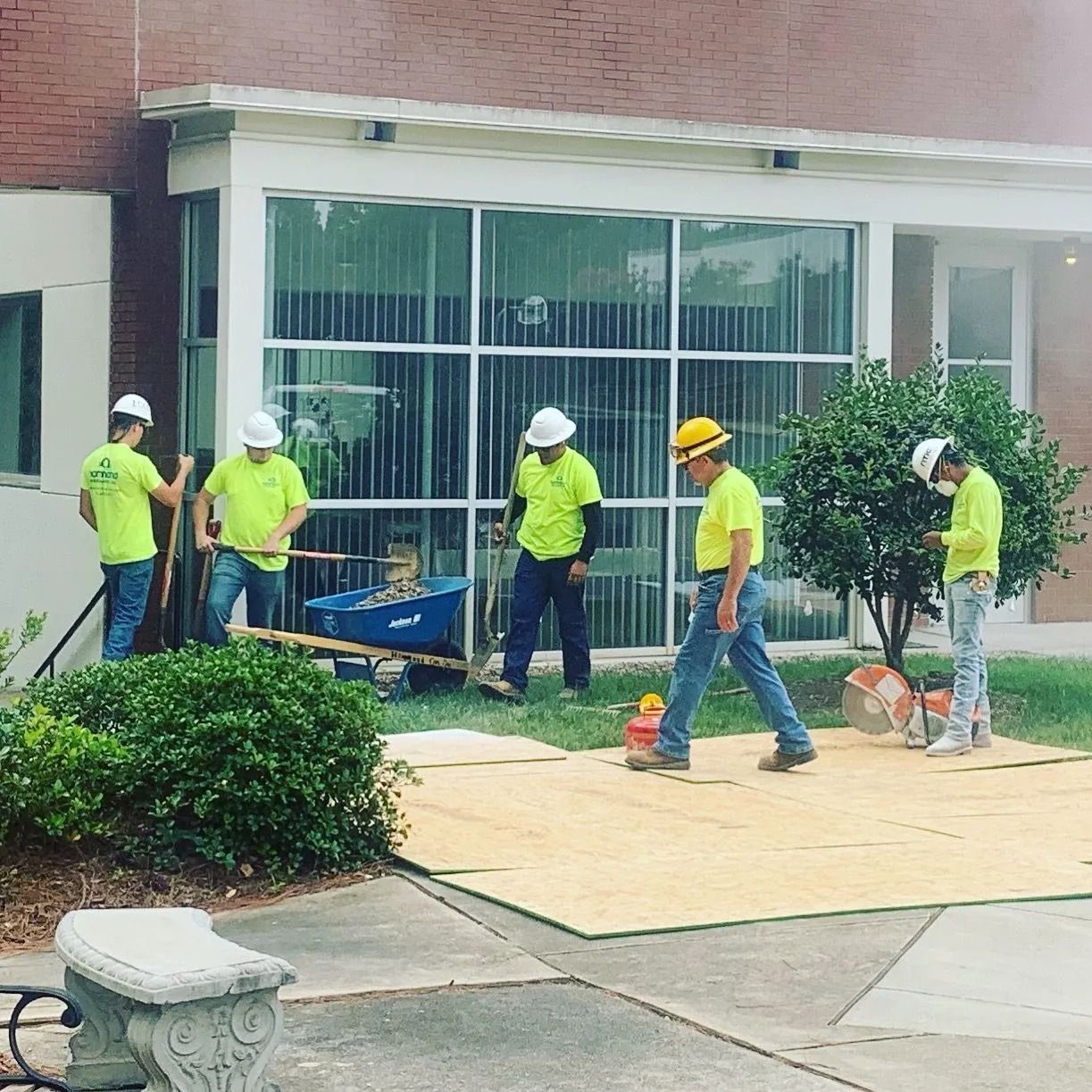 Five construction workers in high-visibility yellow shirts and hard hats work on a plywood platform outside a building.