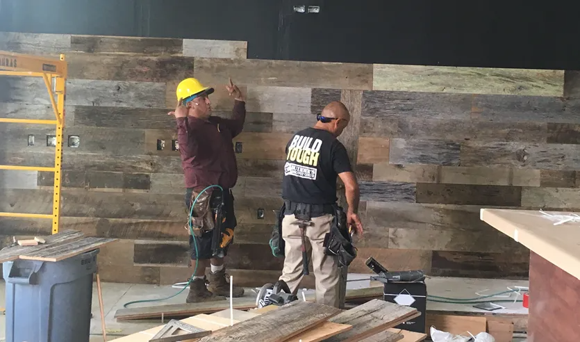 Two workers in hard hats and tool belts install rustic wooden planks on a wall at a construction site.