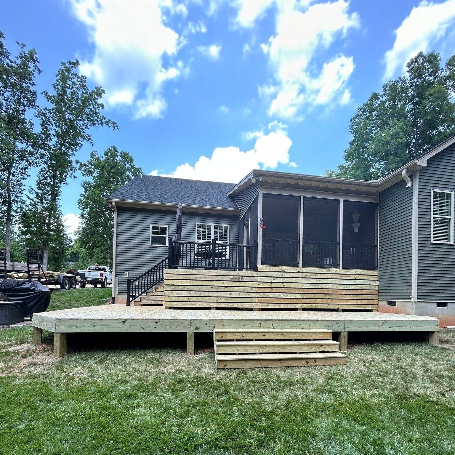 A newly constructed wooden deck featuring wide, stacked steps leads to a screened-in porch on a gray-sided suburban home.
