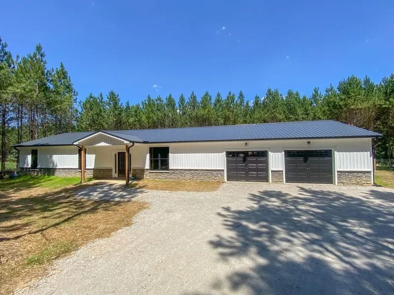 A modern, white single-story building with a dark metal roof, stone foundation, and two garage doors in a wooded setting.
