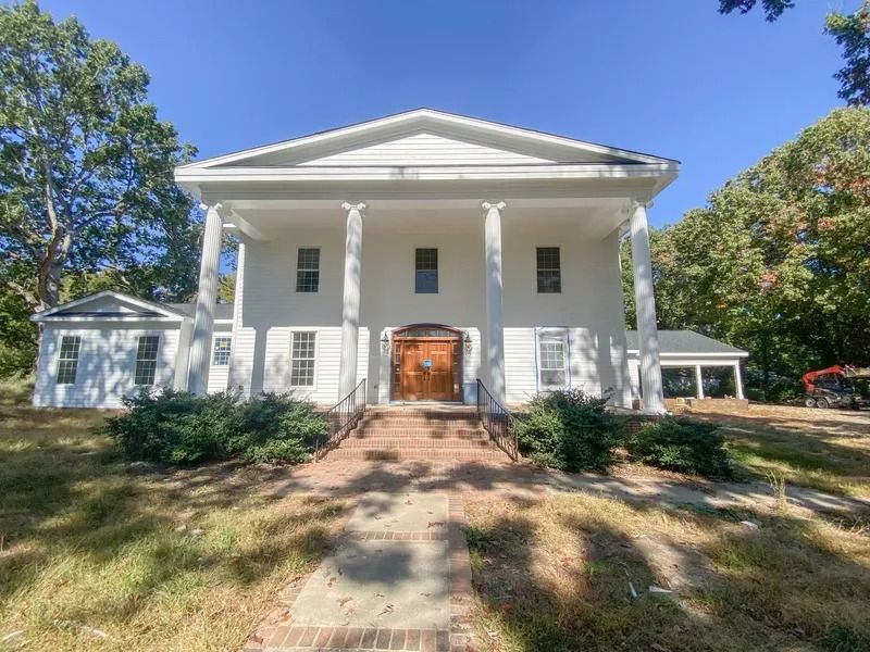A two-story, white classical-style building with a front porch, tall columns, and a brick staircase set among trees.