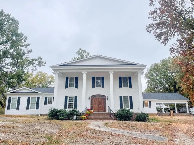 A large, white, two-story colonial-style house with pillars, dark shutters, and a front porch, set among autumn trees.
