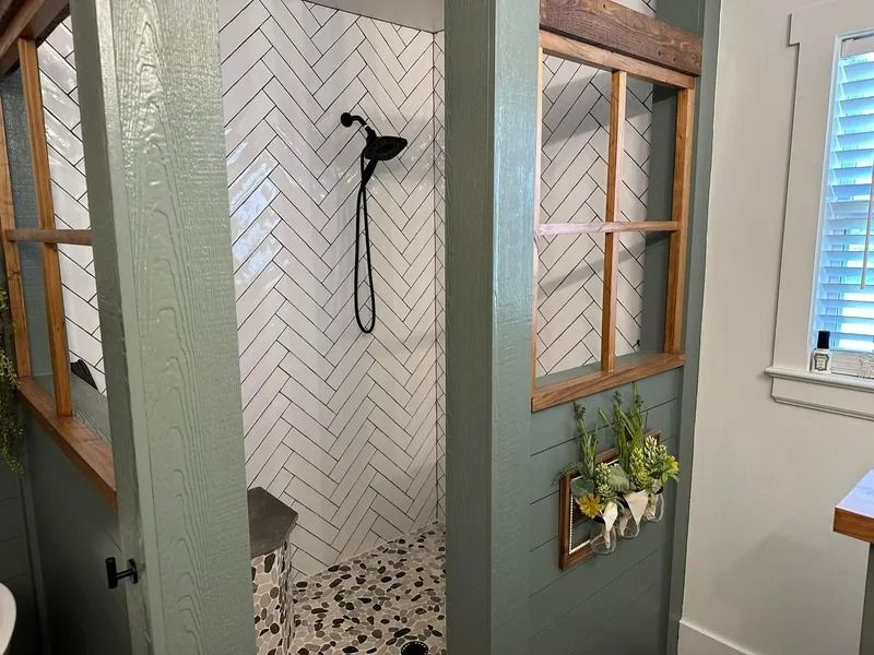 A shower with white herringbone wall tiles and pebble flooring, enclosed by green wooden walls with glass window inserts.