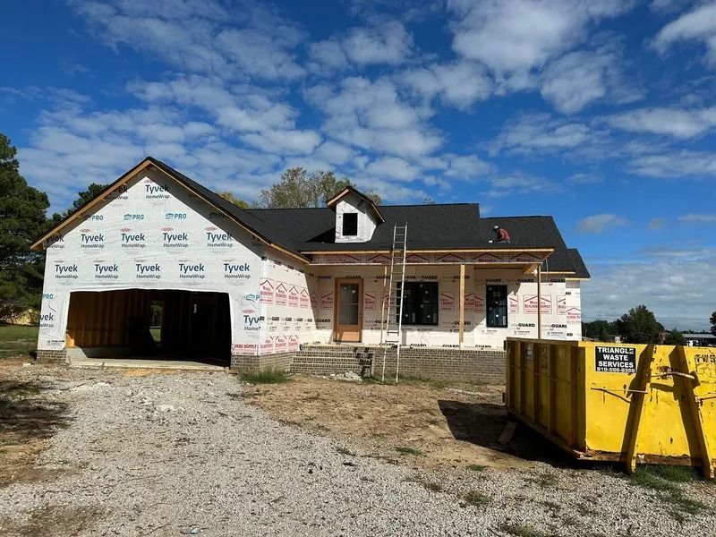 A house under construction with a gravel driveway, a large yellow dumpster, and a blue sky with clouds in the background.