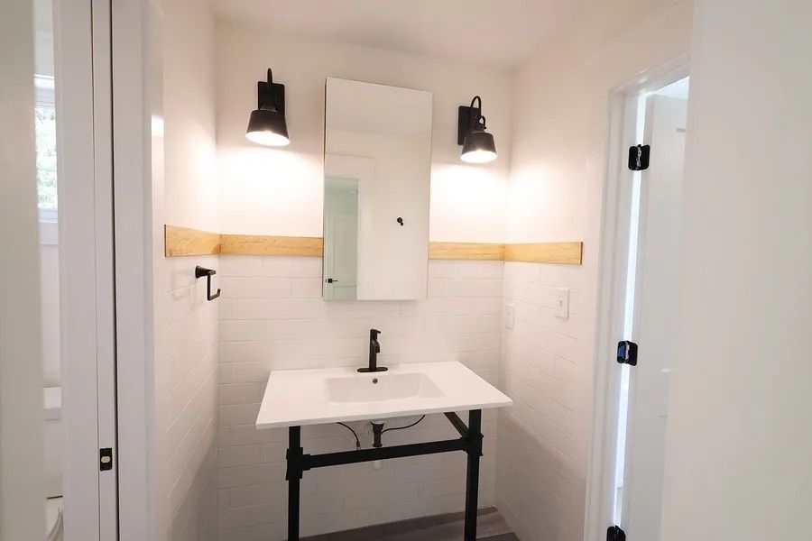 Modern white bathroom with a pedestal sink, black metal frame, two wall sconces, a mirror, and natural wood trim accents.