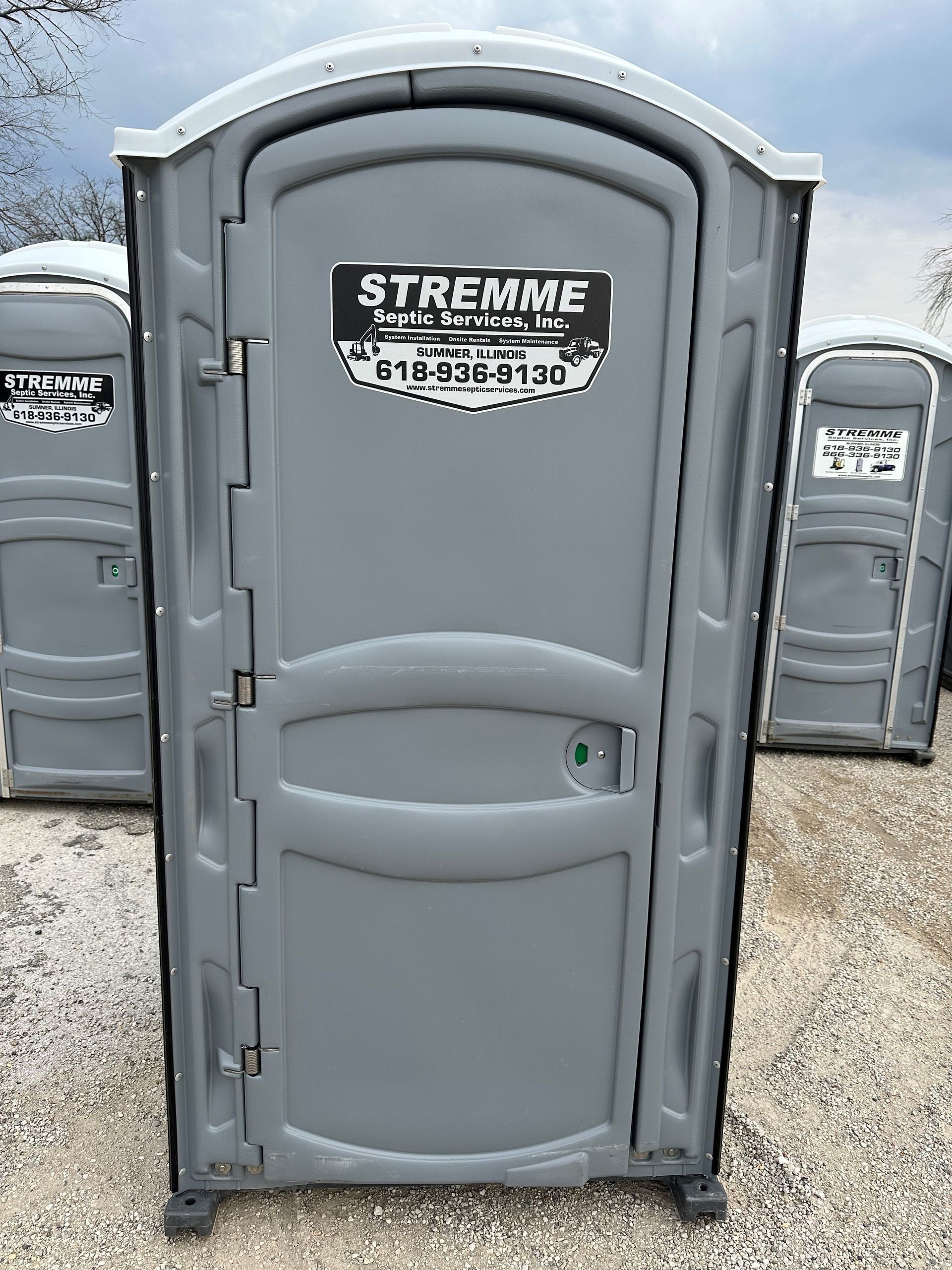 A gray portable toilet is sitting in a gravel lot.