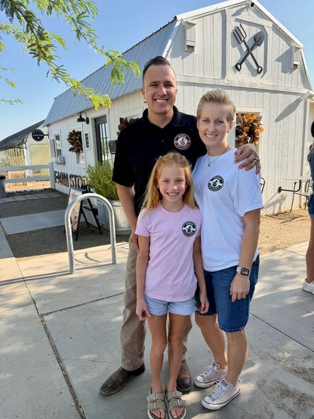 Three people standing outside a white barn, smiling in casual clothes.
