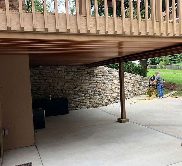 Covered patio with stone retaining wall, concrete floor, and a worker using equipment on the right.