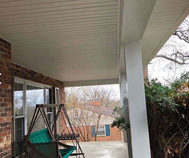 Covered porch with white ceiling, brick wall, swinging chair, and snowy yard beyond a window.