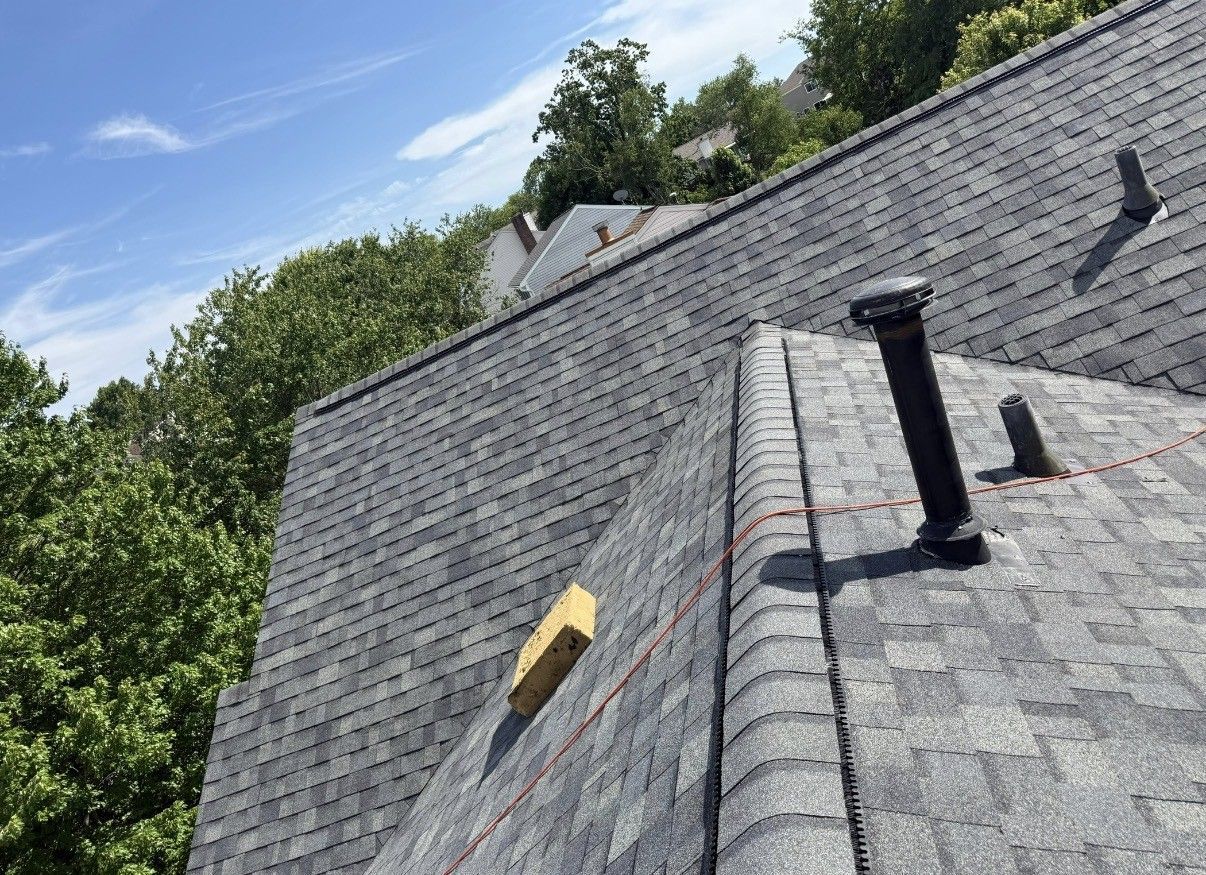 Gray shingled roof with vents and a yellow tool on the slope, trees and blue sky in the background