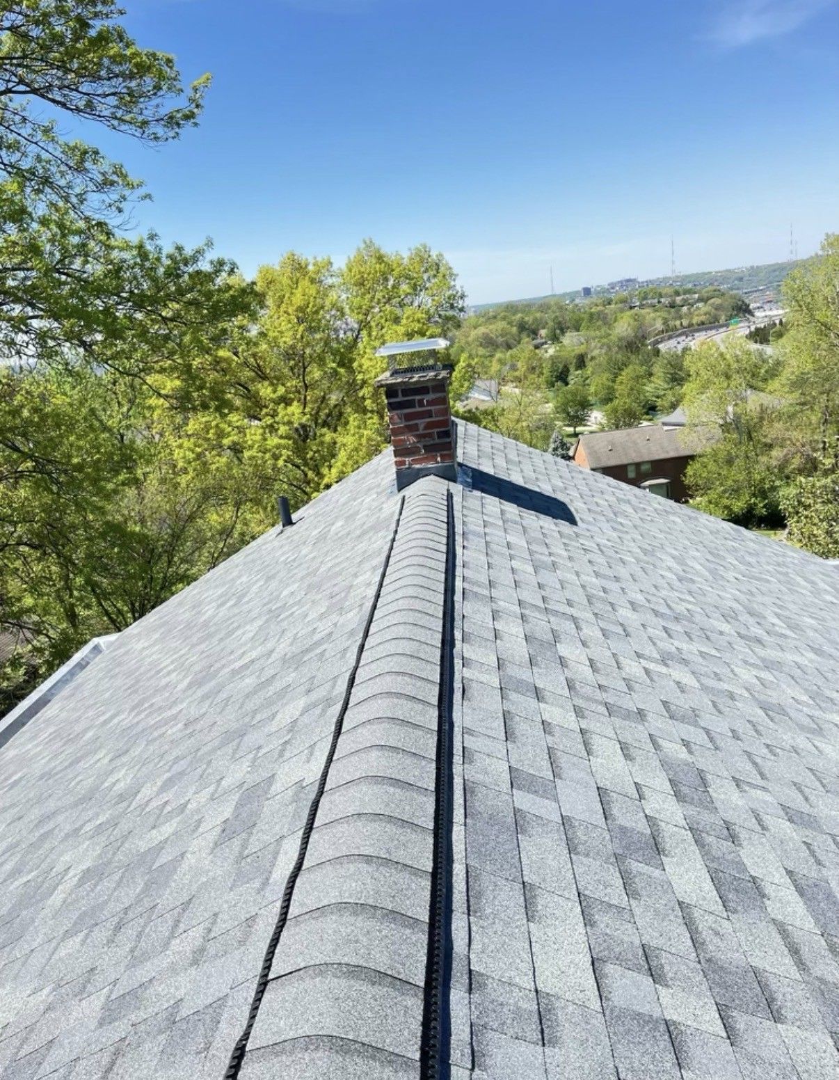 Rooftop with gray shingles and chimney, overlooking green trees under a clear blue sky