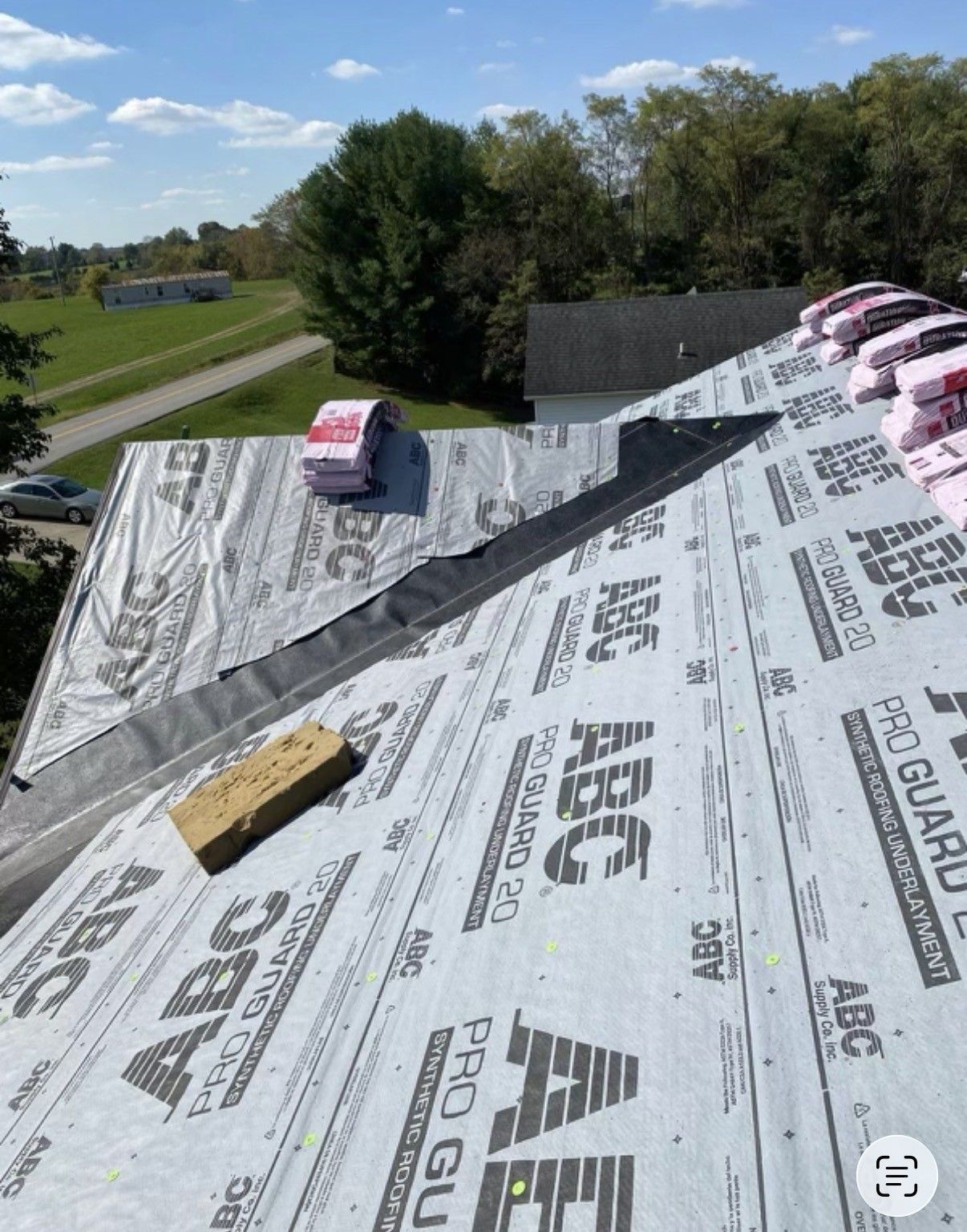 Rooftop underlayment with tools and bundles on a sloped roof, overlooking trees and fields