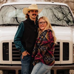 Couple posing in front of white truck; man in cowboy hat, vest; woman in patterned shirt.