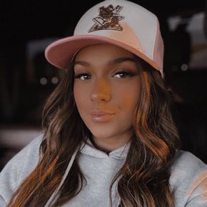 Woman in gray hoodie and pink-and-white trucker hat, looking at the camera. Her brown hair is wavy.