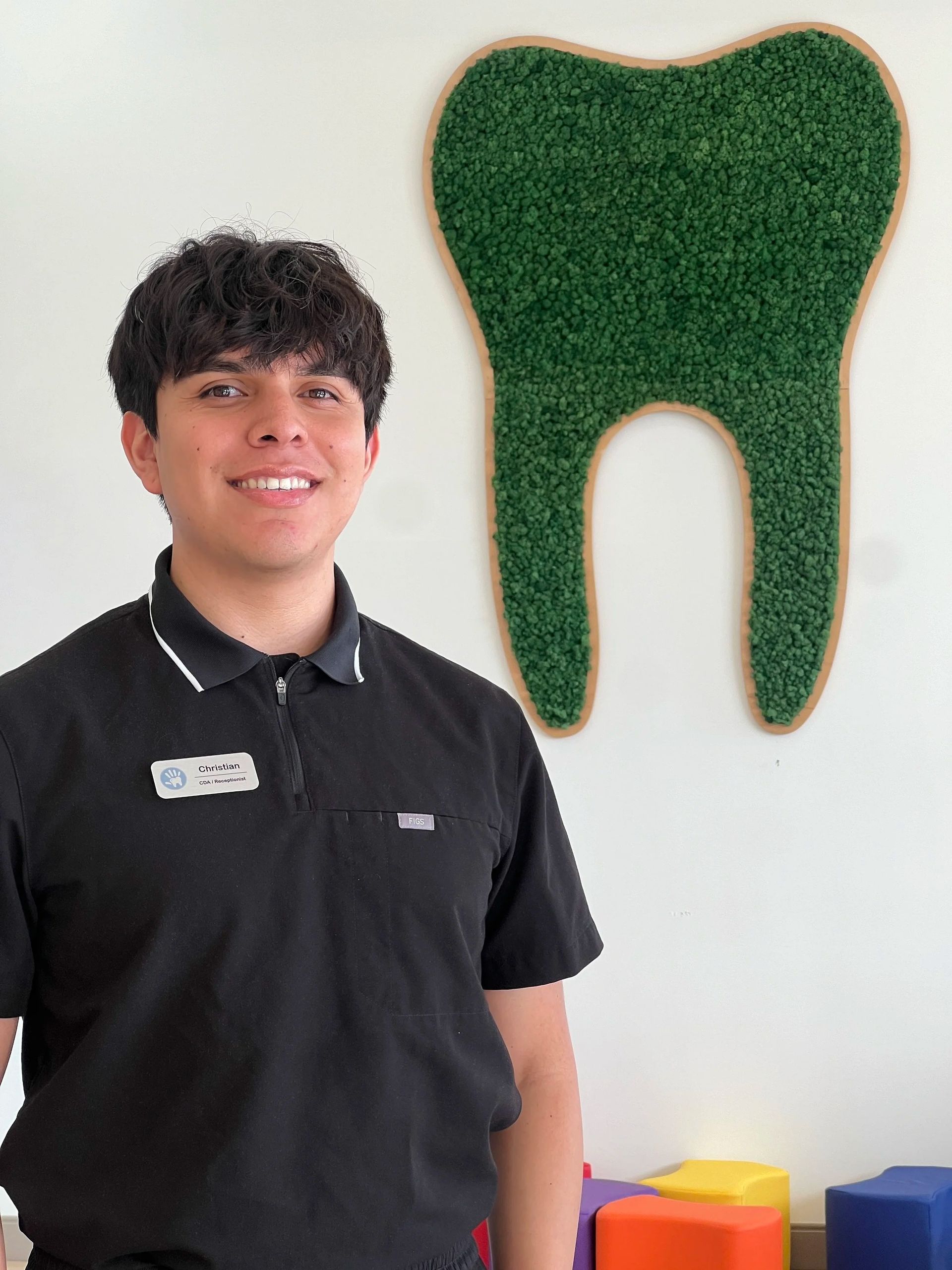 A person in a black polo shirt smiles in front of a wall featuring a green decorative tooth.