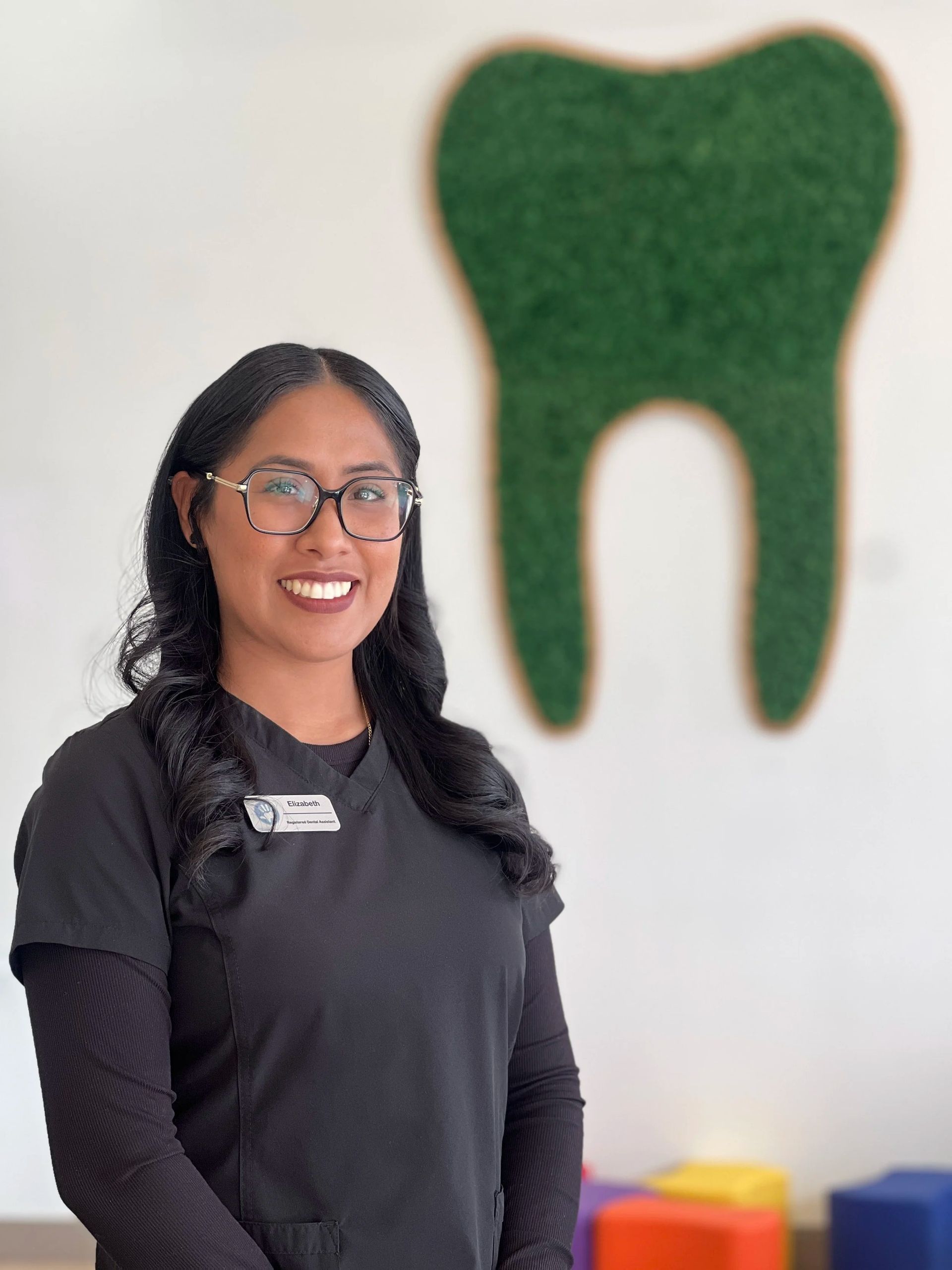 A professional in black scrubs and glasses smiling in front of a wall-mounted green decorative tooth.