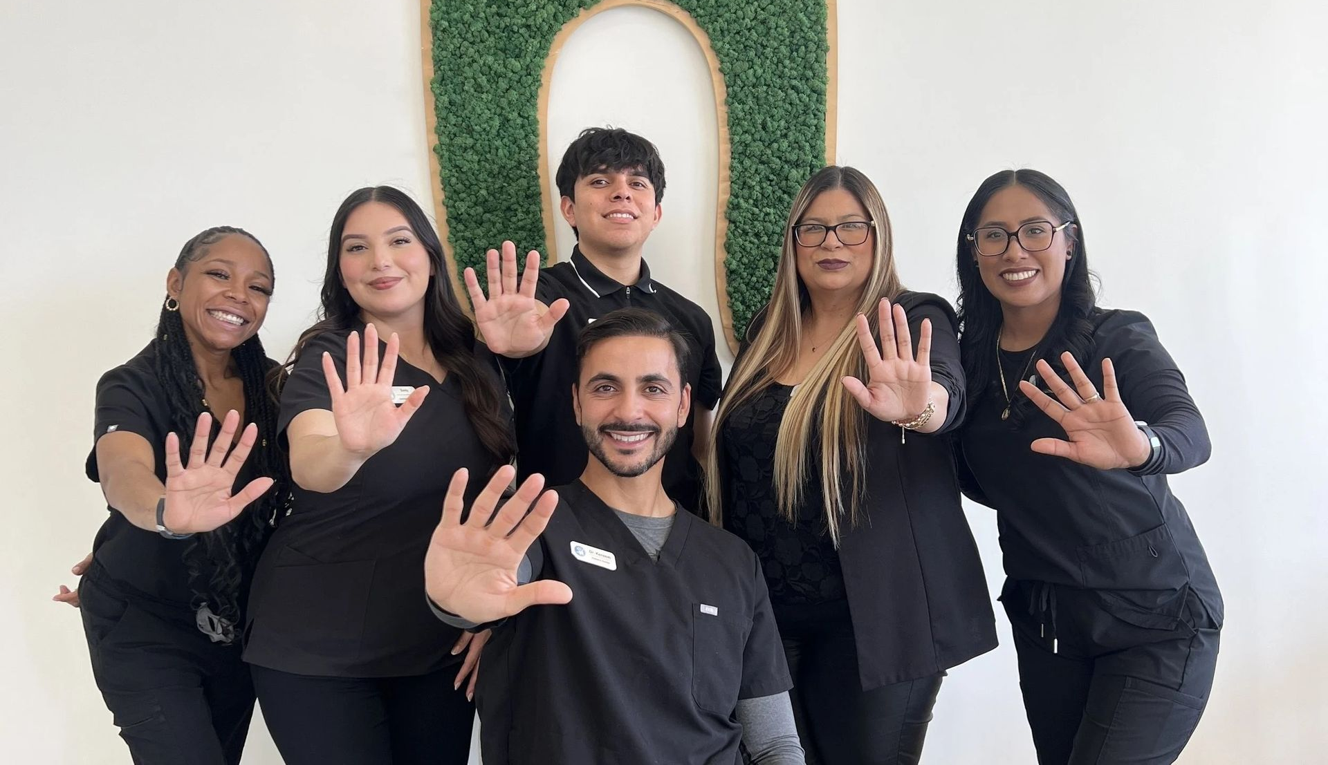 Six people in black uniforms stand in front of a green wall arch, smiling and holding up their hands in a wave.