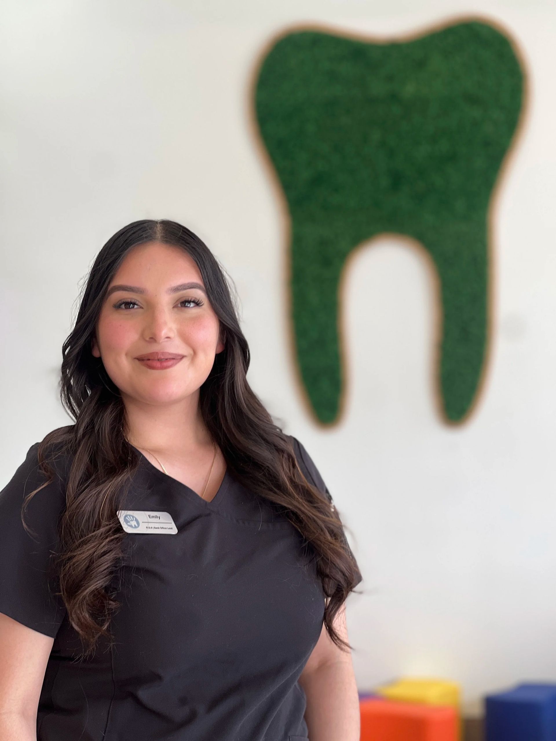A person in a black scrub top with a name tag, posing in a dental office in front of a green tooth-shaped wall decor.