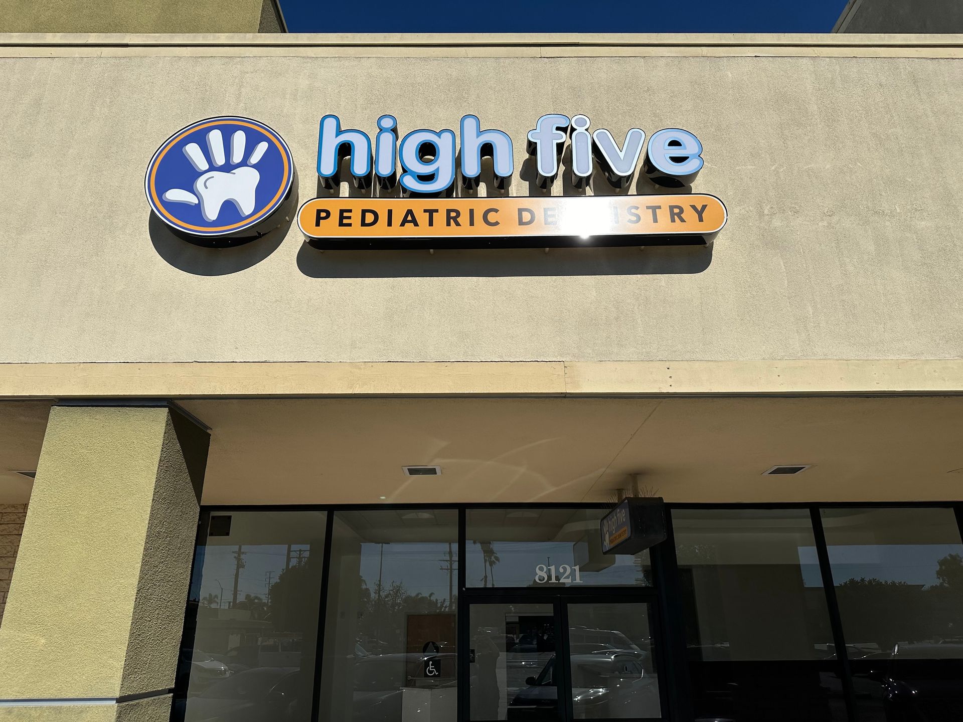 Sign for High Five Pediatric Dentistry with a blue hand-shaped logo containing a white tooth, mounted on a building.