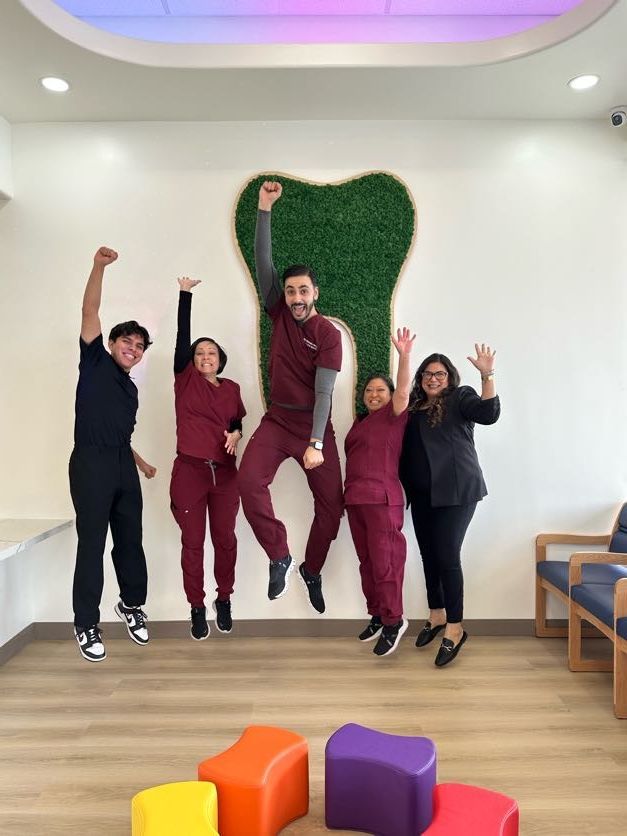 Five dental office staff members jump for joy in front of a green wall with a large, decorative tooth cutout.
