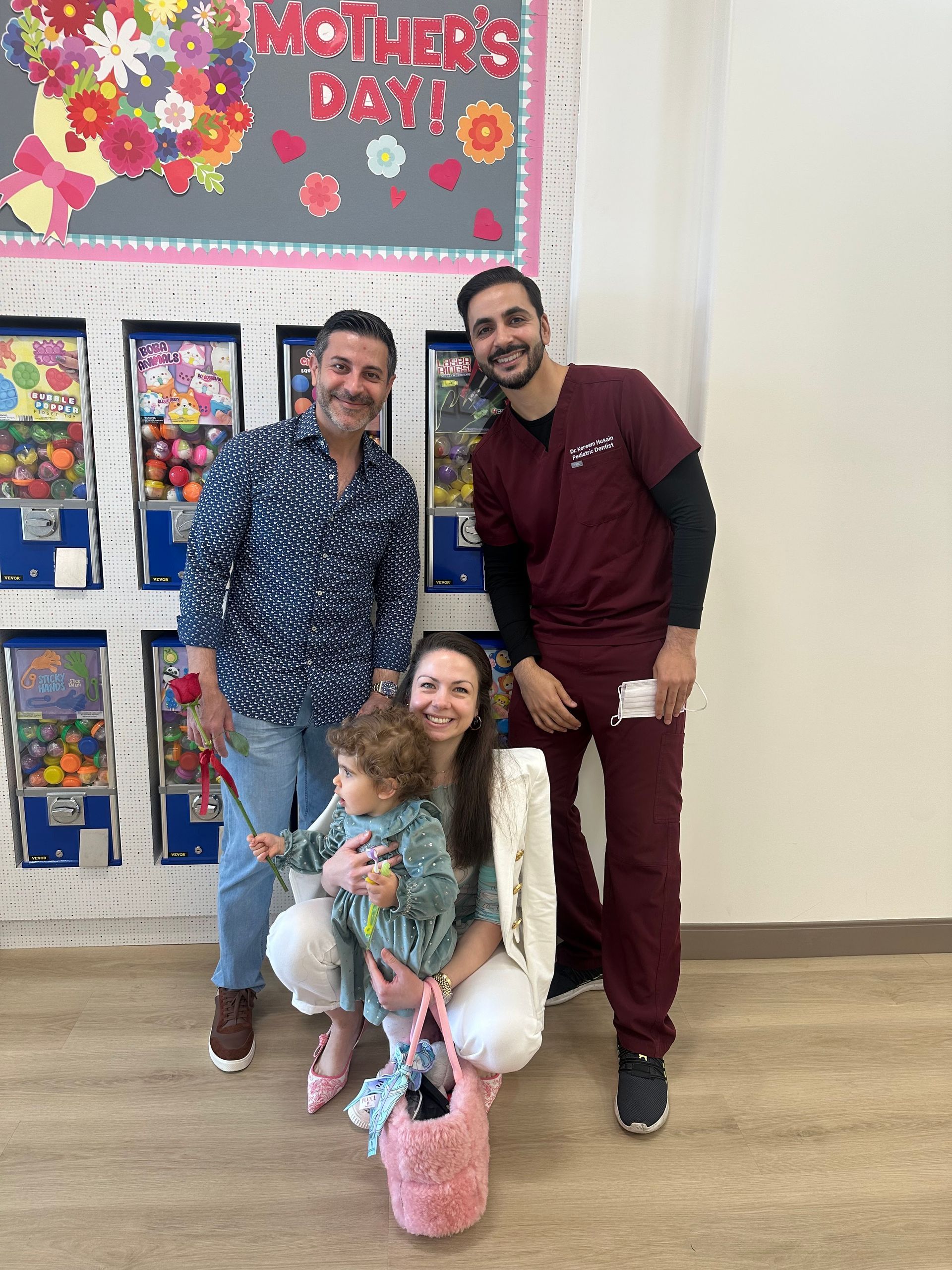 Two men and a woman holding a toddler pose in front of a Mother’s Day bulletin board near vending machines.