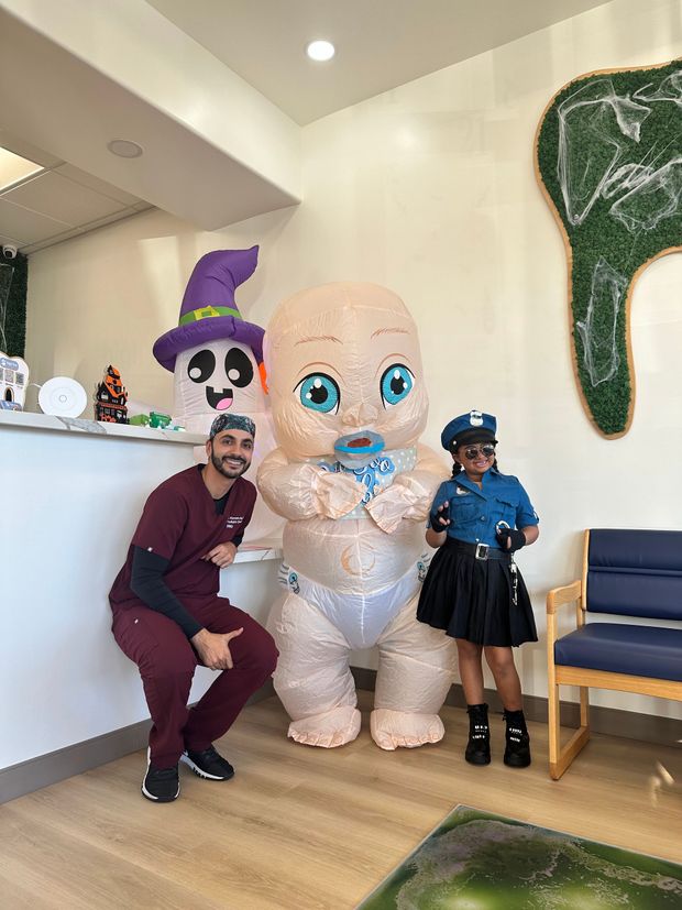 A man in maroon scrubs, a person in a baby costume, and a person in a police costume pose together in an office.