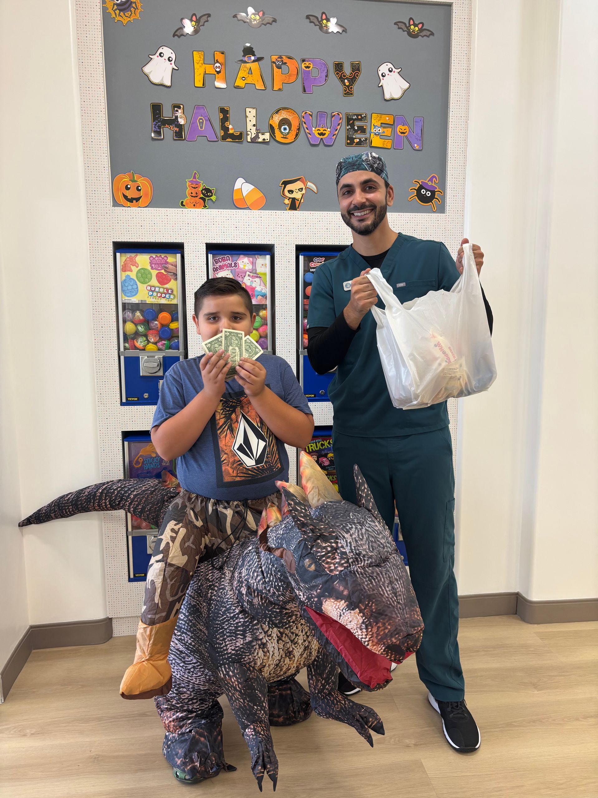 A person in a dinosaur costume and an adult in scrubs holding a bag stand in front of a Halloween-decorated wall.