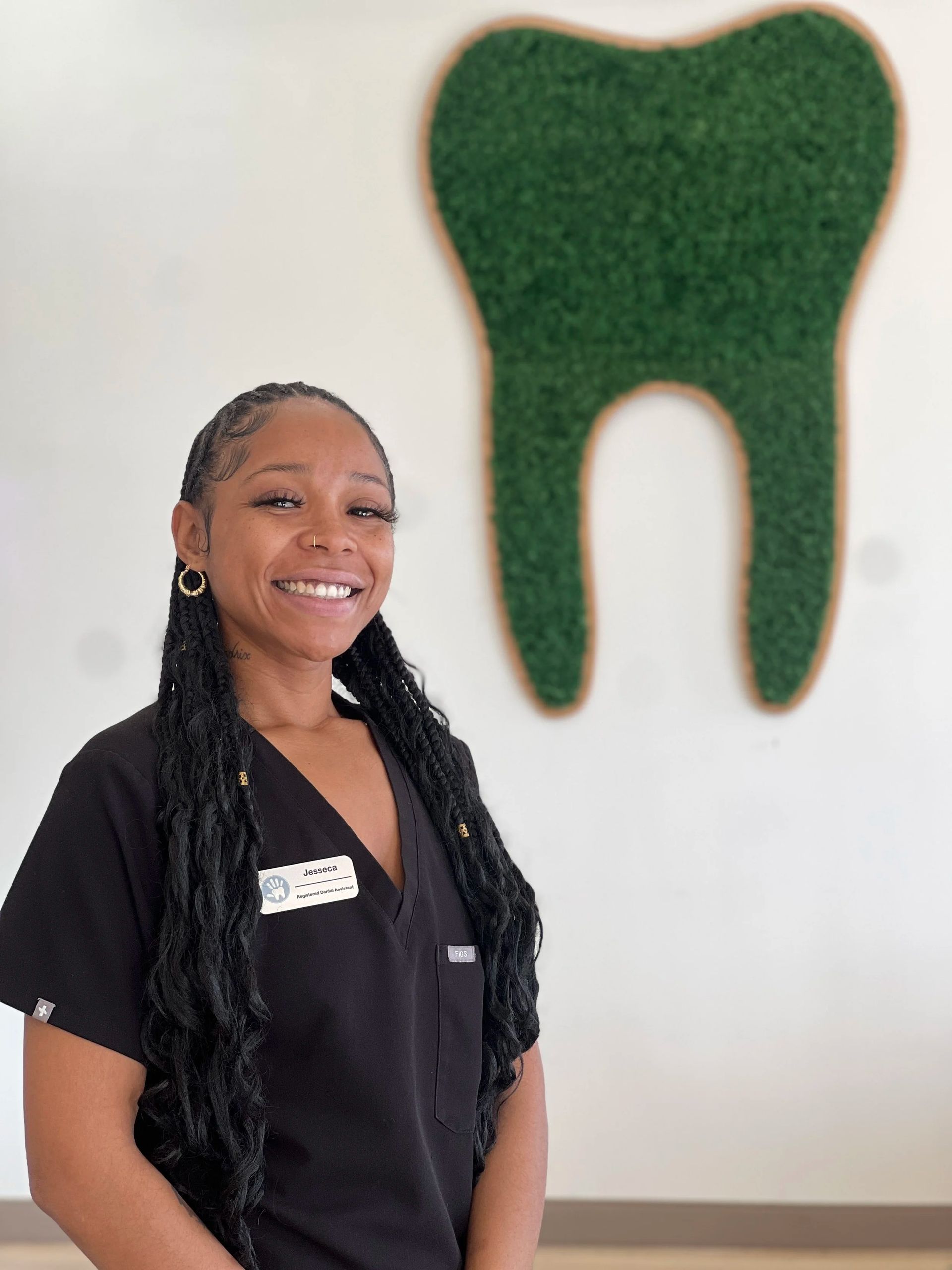 A smiling person wearing black scrubs stands in front of a wall featuring a large, green, moss-covered tooth decoration.