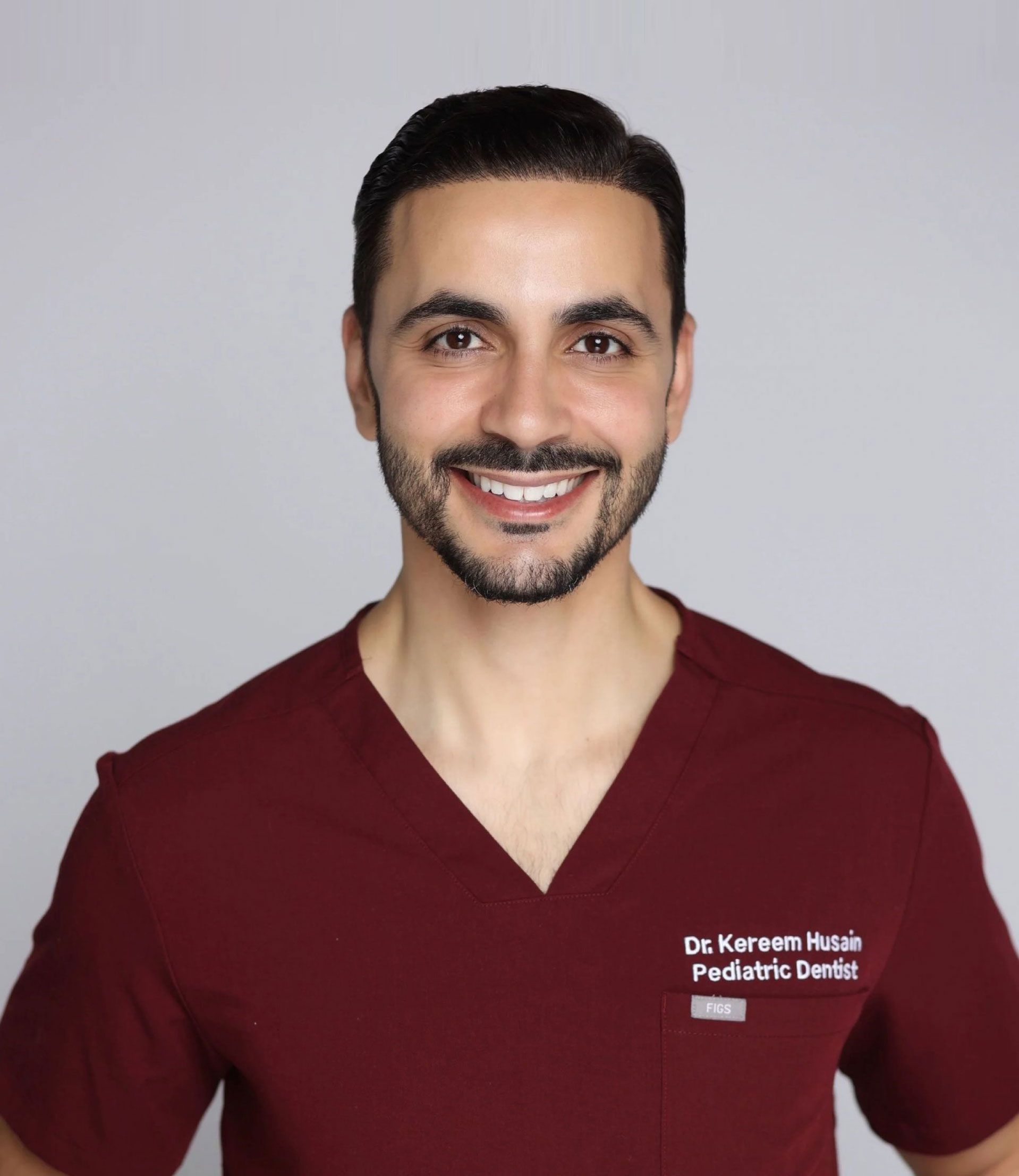 A person in a maroon scrub top smiling against a light gray background, with text on their chest reading