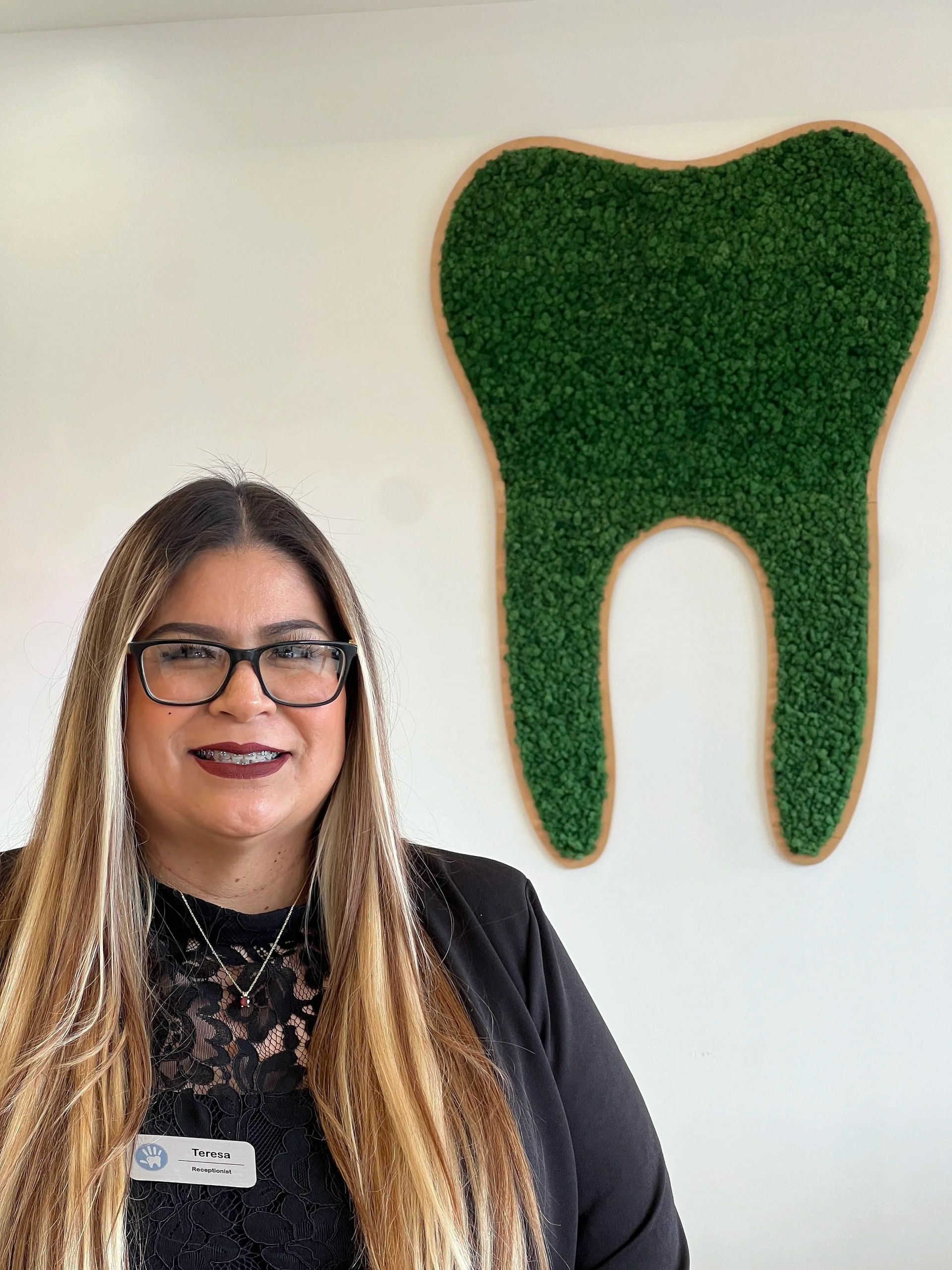 A smiling professional wearing glasses stands in front of a wall-mounted, green moss-covered tooth logo.