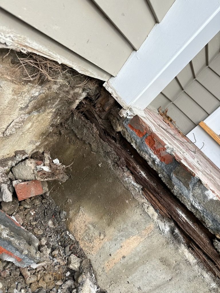 Damaged house corner with exposed foundation, broken concrete, and scattered debris near siding and brick trim