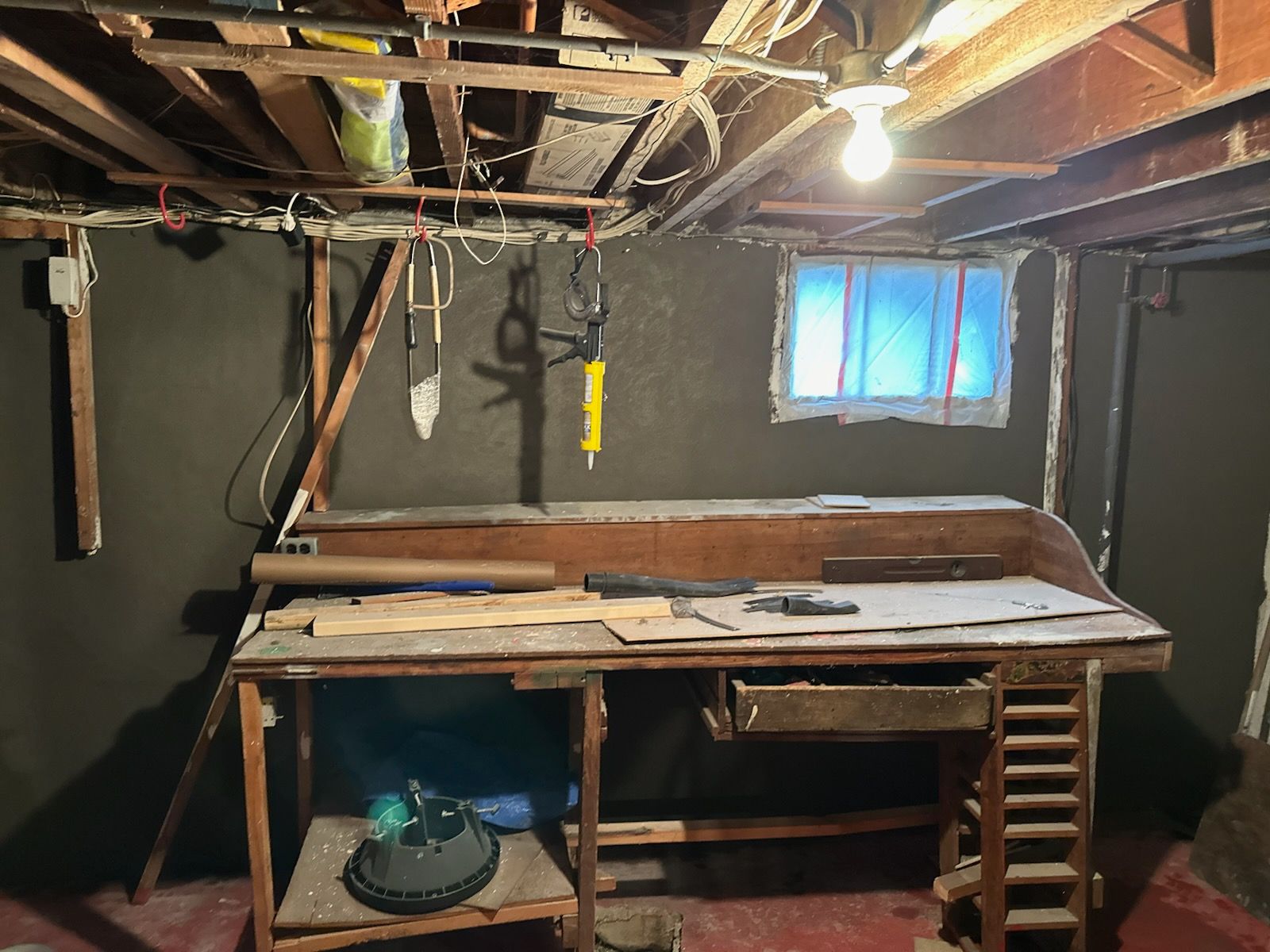 Dim basement workspace with a wooden bench, hanging light, exposed beams, and a small window.