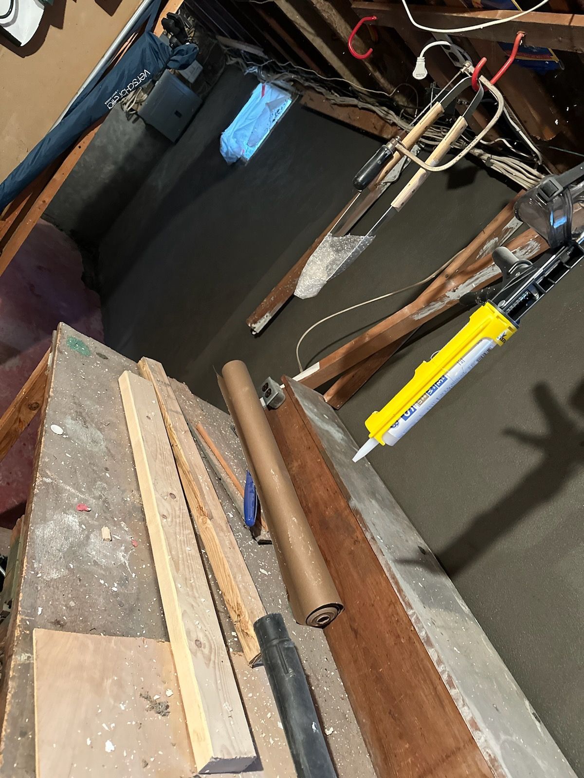 Cluttered workshop bench with wood planks, pipes, and hanging tools against a dark wall.
