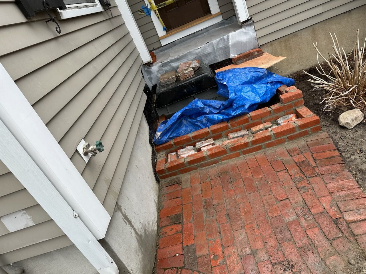 Blue tarp covering items on a brick patio beside a house, with a grill and steps nearby.