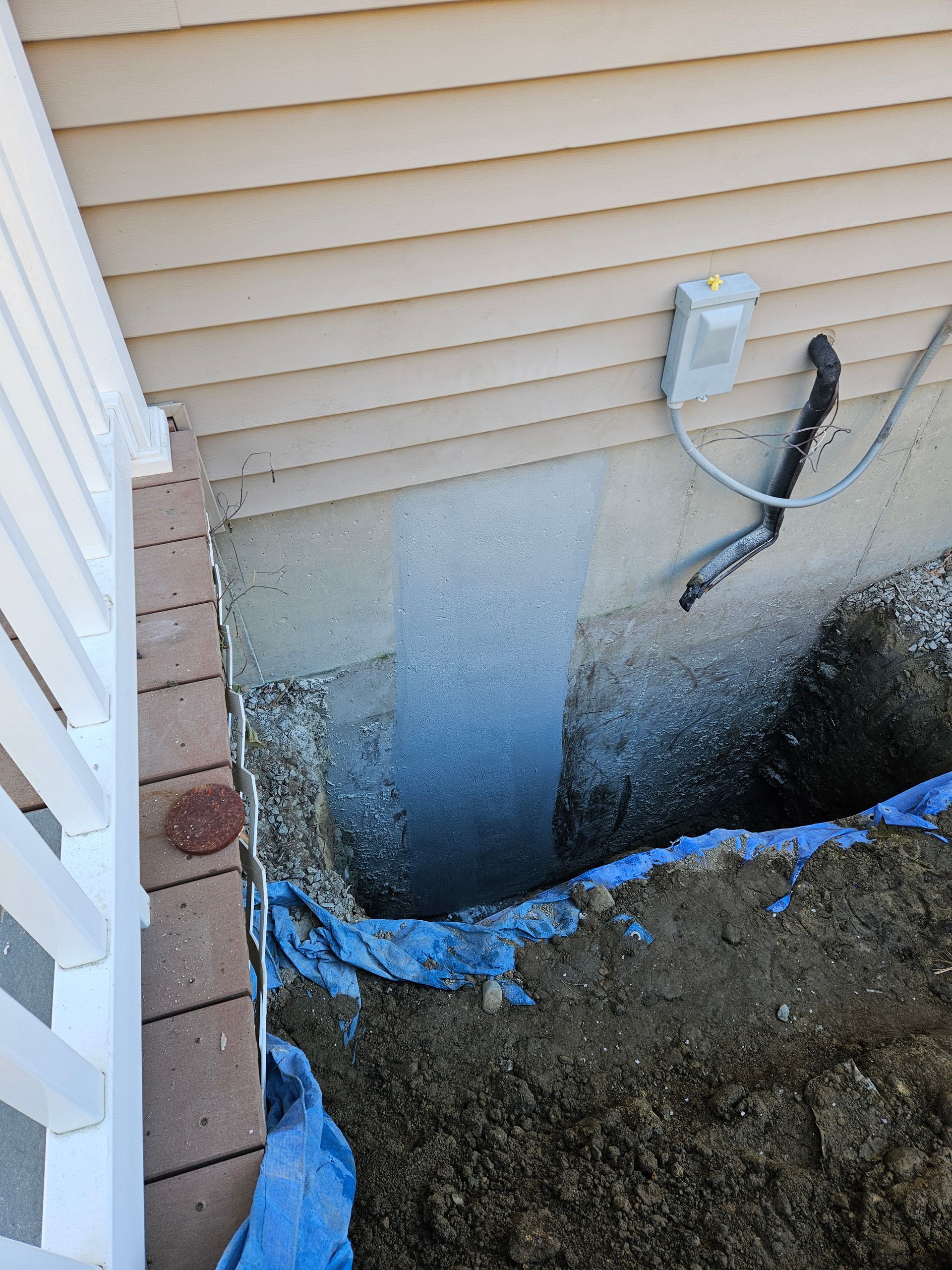 Excavated hole beside house foundation with blue tarp, exposed wall, and utility meter on siding