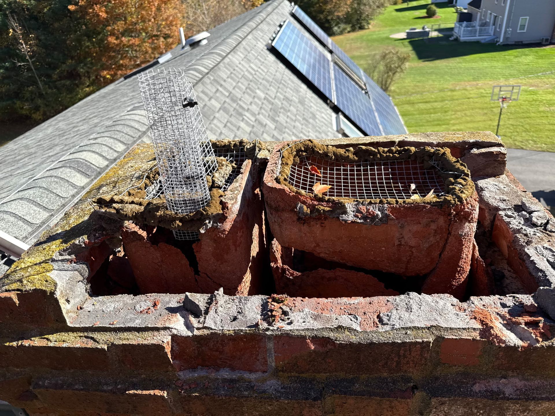 Damaged brick chimney with two broken flue openings on a sloped roof with solar panels.